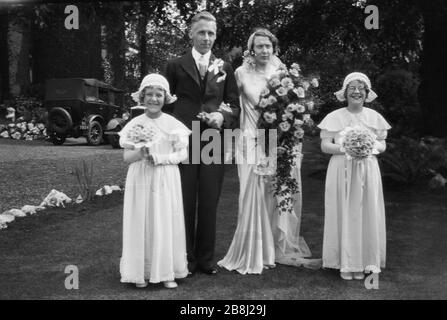 Anni '20, storico, tenendo i loro mazzi di fiori, una sposa e lo sposo in piedi fuori per una foto, con le sue bridesmaids, due ragazze, in moda del giorno, con automobili dell'epoca parcheggiata sul viale, Inghilterra, Regno Unito. Foto Stock