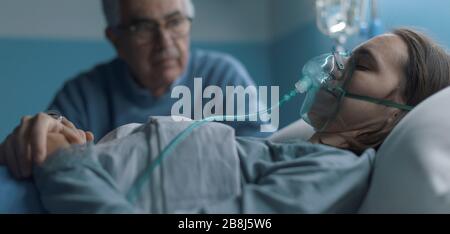 Uomo anziano interessato che assiste la sua figlia giovane che giace nel letto all'ospedale, sta tenendo la sua mano e seduta accanto a lei Foto Stock