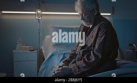Uomo anziano solitario seduto sul letto dell'ospedale di notte e tenendo una bottiglia di pillola, soffre di insonnia e si sente depresso Foto Stock