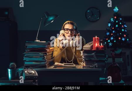 Infelice donna d'affari stressata che lavora a tarda notte in ufficio la vigilia di Natale, sta tenendo due ricevitori telefonici e rispondendo alle telefonate Foto Stock