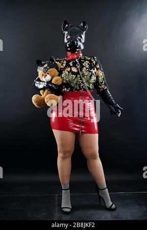 donna più taglia con maschera in gomma per la protezione posando con il suo orso peloso su fondo nero da solo. Foto Stock