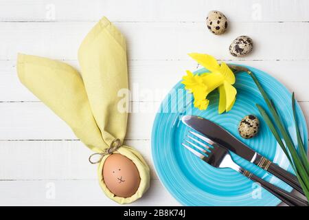 Bella festa tavola di Pasqua impostazione con uovo in tovagliolo come coniglietto di Pasqua e fiori di primavera su tavolo di legno bianco. Vista dall'alto con spazio di copia. Foto Stock