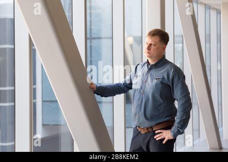 Un uomo di 40-45-anni in una camicia blu a quadri si trova nel corridoio posteriore dell'ufficio in mezzo a una grande finestra. Foto Stock