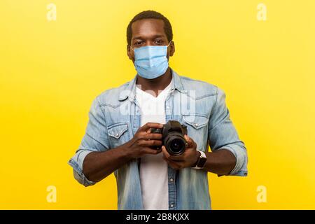 Ritratto di fotografo positivo, uomo con maschera medica in possesso di fotocamera reflex digitale professionale e guardando con un sorriso toothy, godendo il suo lavoro. ind Foto Stock