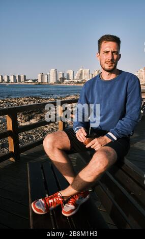Un bell'uomo che beve l'infusione di Yerba Mate nella rambla di Punta del Este, Maldonado, Uruguay con l'oceano atlantico e la città sullo sfondo Foto Stock
