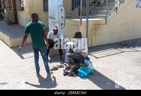 Zwelihle, Hermanus, Capo Occidentale, Sudafrica. Uomo rammendo e pulendo scarpe sulla strada a bordo strada con gli amici. Foto Stock