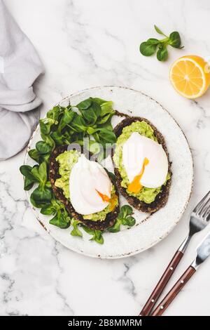 Toast di segale con avocado schiacciato, foglia di insalata verde e uovo in camicia sul piatto, sfondo del tavolo in marmo Foto Stock