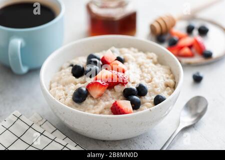 Porridge d'avena con frutti di bosco, miele e tazza di caffè sul tavolo bianco. Cibo sano per la colazione, piatto di avena vegetariana porridge Foto Stock