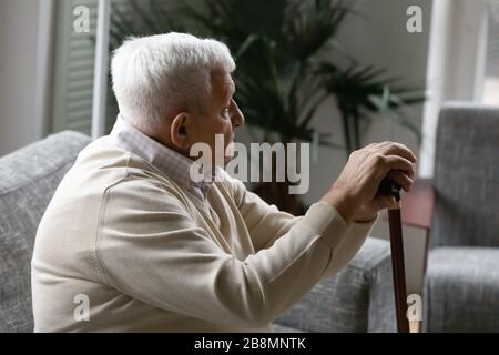 L'uomo anziano siede sul divano tenendo il bastone da passeggio si sente solo Foto Stock