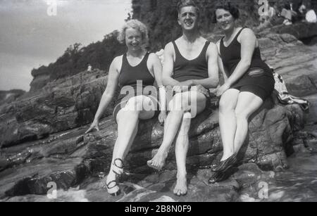 Alla fine degli anni '20, all'inizio degli anni '30, un uomo e due Signore seduti insieme su alcune rocce sulla costa nei costumi da bagno dell'epoca. Foto Stock