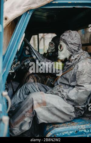 Due persone siedono in tute protettive NBC e maschere a gas nel vecchio camion. Concetto di misure preventive e di protezione per il coronavirus COVID 19 pandemico Foto Stock