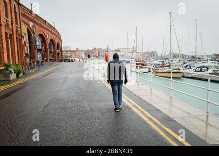 Un uomo con camminate da solo lungo un fronte del porto. La strada ha due linee gialle: Il tempo è umido, indossa un cappotto e un cappello di lana. Potrebbe essere un con Foto Stock