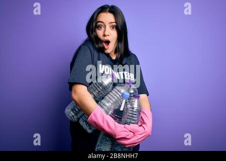 Giovane donna bruna che indossa la t-shirt volontaria raccolta di bottiglie di plastica su sfondo viola spaventato in shock con un viso a sorpresa, impaurito ed eccitato Foto Stock