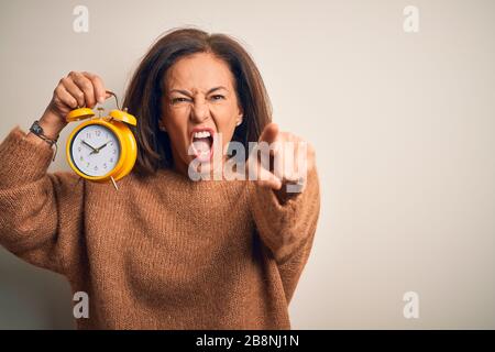 Donna brunetta di mezza età che tiene clasic sveglia su sfondo isolato puntando dispiaciuto e frustrato alla macchina fotografica, arrabbiato e furioso con Foto Stock