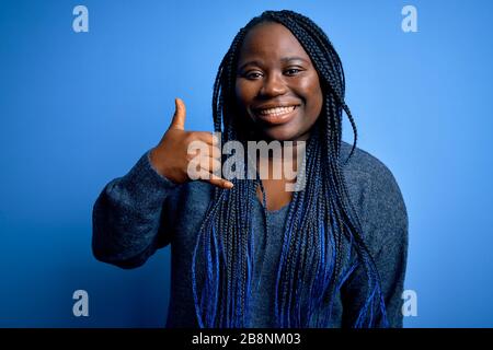 African american Plus taglia donna con trecce indossare maglione casual su sfondo blu sorridendo facendo gesto telefono con mano e dita come talkin Foto Stock