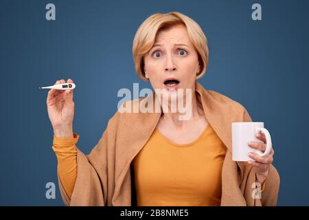 Donna di mezza età che tiene in mano bevande e termometro elettronico Foto Stock