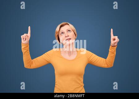 Stile libero. Donna matura in piedi isolata su blu che punta verso l'alto allo spazio di copia interessato Foto Stock
