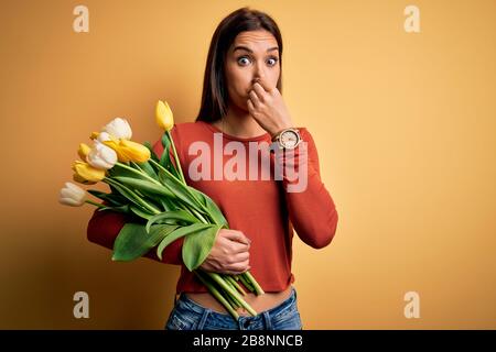 Giovane bella donna brunetta tenuta bouquet di tulipani fiori su sfondo giallo odore qualcosa di stinky e disgustoso, odore intollerabile, h Foto Stock