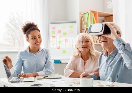 Agente di viaggio in ufficio che utilizza un computer portatile che guarda i clienti senior che indossano cuffie per realtà virtuale sorridenti e entusiasti Foto Stock