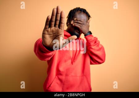 Giovane uomo sportivo afro-americano che indossa felpa con cappuccio su sfondo giallo coprendo gli occhi con le mani e facendo fermare il gesto con Sad e F. Foto Stock