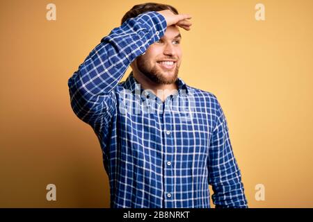 Giovane uomo d'affari biondo con barba e occhi blu indossando camicia su sfondo giallo molto felice e sorridente guardando lontano con mano sopra la testa. Mare Foto Stock
