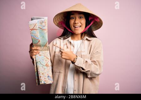 Giovane donna turistica tailandese in vacanza indossando tradizionale cappello asiatico conico tenendo mappa della città molto felice indicando con mano e dito Foto Stock