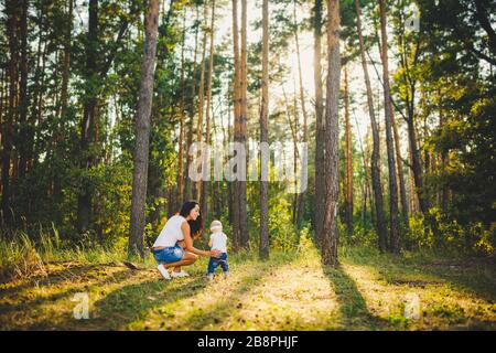 bella ed elegante madre conduce la mano di una ragazza di un anno dalla nascita nel parco contro lo sfondo di alti alberi di conifere. Famiglia Foto Stock