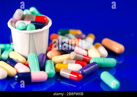 Varie capsule colorate assortite con tazza di carta medicazione su superficie riflettente blu. Foto Stock