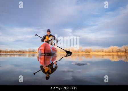 Un adulto pagatore maschio in una muta sta remando un paddleboard stand up su un lago tranquillo in Colorado, inverno o primi paesaggi primaverili, camer bassa angolazione Foto Stock