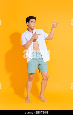 Corpo pieno di giovane bell'uomo asiatico in un outfit estivo casual che punta due mani da parte su sfondo giallo colorato Foto Stock