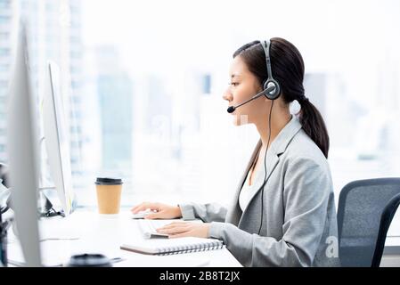 Giovane donna d'affari asiatica che indossa cuffie e auricolari che lavorano nell'ufficio del call center come operatore di telemarketing Foto Stock