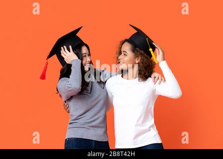 Ritratto di due donne happy misto-corsa che si laurea tenendo lì tappi di graduazione guardando l'altro in arancione isolato sfondo studio Foto Stock