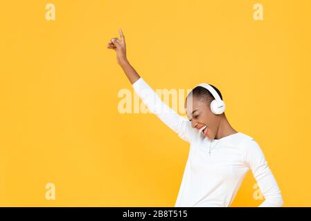 Felice giovane African American donna che alza mano in su mentre ascolta la musica in cuffie senza fili su sfondo studio isolato giallo Foto Stock