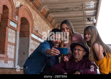 Uomo paralitico caucasico in carrozzina accompagnato da un giovane latino e da una ragazza caucasica che scattano una foto con il suo cellulare felice sulla strada Foto Stock