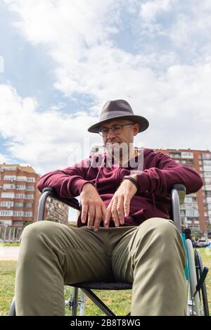 Uomo caucasico con occhiali e cappello depressivo e triste in una sedia a rotelle solitaria in un parco con il cielo e alcuni edifici sullo sfondo con lo spazio Foto Stock