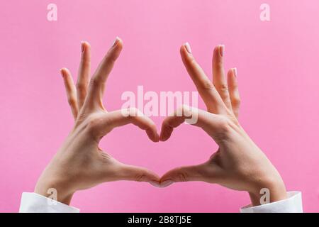 Mani femminili che formano un simbolo del cuore su sfondo rosa Foto Stock