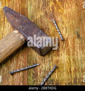 Vecchio martello arrugginito e tre chiodi in acciaio giacciono su una tavola di legno graffiata. Spazio di copia per il testo. Messa a fuoco selettiva. Concetto di lavoro domestico. Formato quadrato. Chiudi- Foto Stock