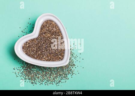 Semi interi di chia grezzi in un piatto a forma di cuore su sfondo blu Foto Stock