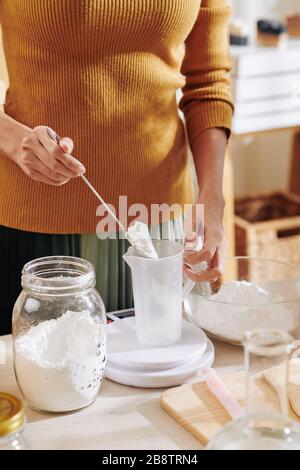 Immagine tagliata della donna che mette la polvere di liscivia di soda in vaso di plastica e la misura su scala elettronica quando fa la miscela del sapone Foto Stock