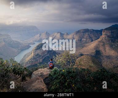 Uomo caucasico seduto sul bordo della scogliera ammirando la vista mozzafiato di 3 rondavel dal punto elevato a Mpumalanga Sud Africa percorso panoramico Foto Stock