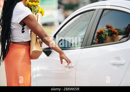 Immagine tagliata della donna con borsa della spesa e bouquet di girasoli che aprono la porta dell'auto Foto Stock