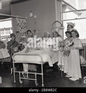 Anni '60, storico, una visita in un ospedale da parte della recentemente incoronata London May Queen, una giovane adolescente e entourage, un gruppo di bambini piccoli vestiti in costumi, Lewisham, South East London, Inghilterra, Regno Unito. I bambini siedono accanto ad un paziente anziano di sesso maschile seduto nel suo letto dopo avergli presentato una torta di compleanno. Foto Stock