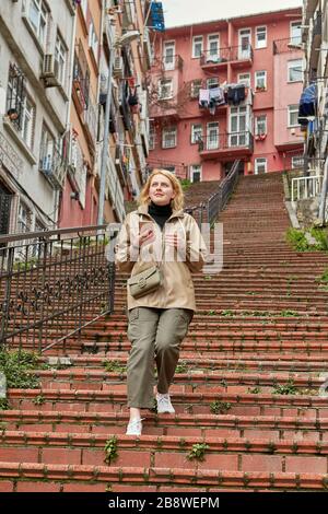 Una giovane donna bianca in abiti invernali caldi scende i gradini di una ripida scala rossa a Istanbul, e tiene un telefono cellulare con un navigatore e un ma Foto Stock