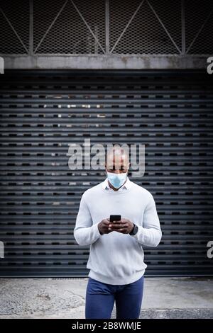 African American che indossa la maschera coronavirus covid19 e che usa il suo telefono per strada Foto Stock