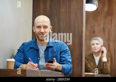 Ritratto di uomo calvo adulto sorridente felicemente alla macchina fotografica mentre lavorando al tavolo del caffè e tenendo smartphone, spazio copia Foto Stock