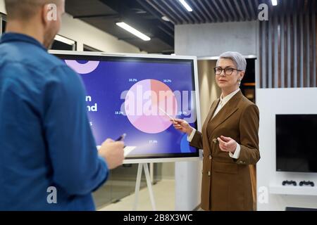 Ritratto di moderna donna d'affari matura che punta al grafico a torta mentre dà la presentazione per i dipendenti o i clienti nella sala d'ufficio, spazio di copia Foto Stock