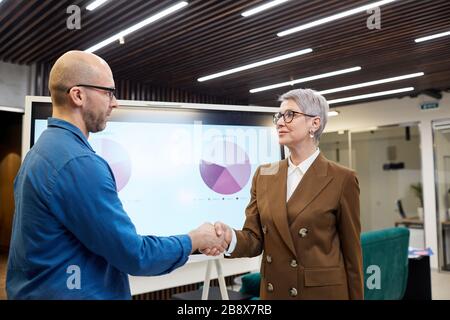 Girovita ritratto di donna maturo manager scuotendo le mani con il cliente o dipendente mentre in piedi contro il bordo di presentazione, spazio copia Foto Stock