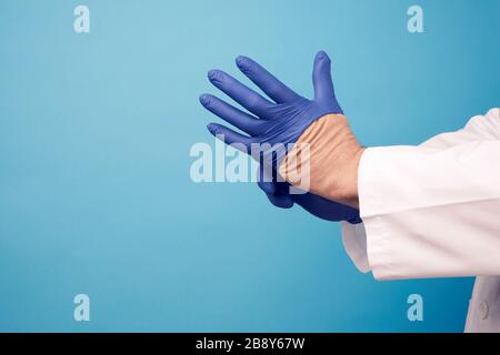 medico maschile in uniforme bianca mette sulle sue mani guanti in lattice blu sterile prima dell'intervento chirurgico, sfondo blu, spazio copia Foto Stock