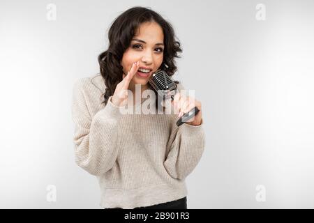 la ragazza bruna in un maglione bianco tiene un microfono retrò in mano e canta una canzone su uno sfondo bianco Foto Stock