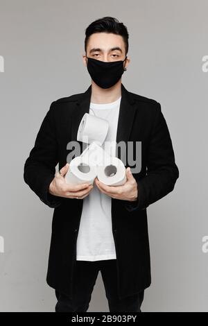 Bel giovanotto in maschera nera protettiva con quattro rotoli di carta igienica, isolato su sfondo grigio. Giovane uomo in maschera protettiva del viso Foto Stock
